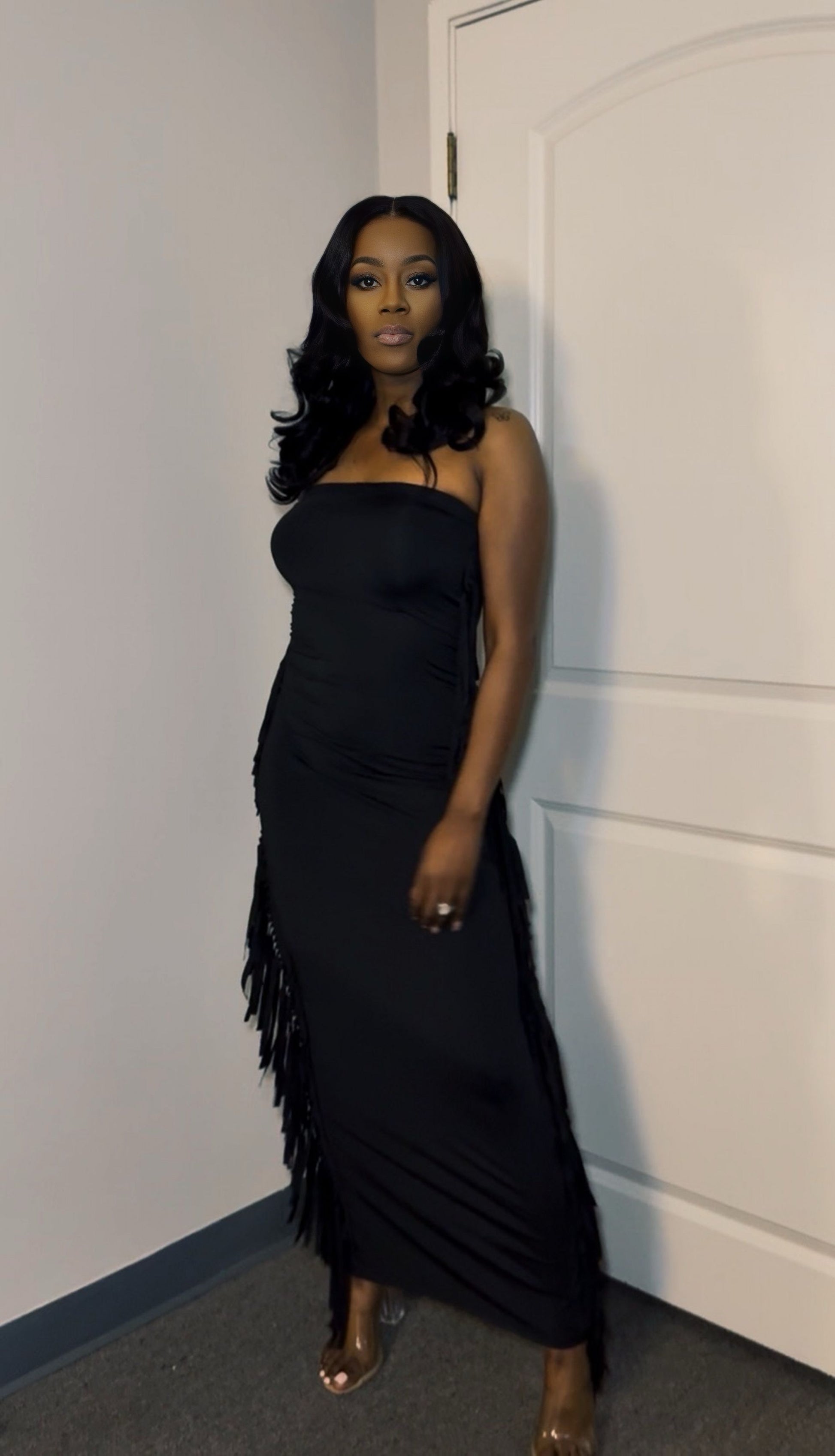 Woman in a black strapless dress with fringe details standing against a white wall.