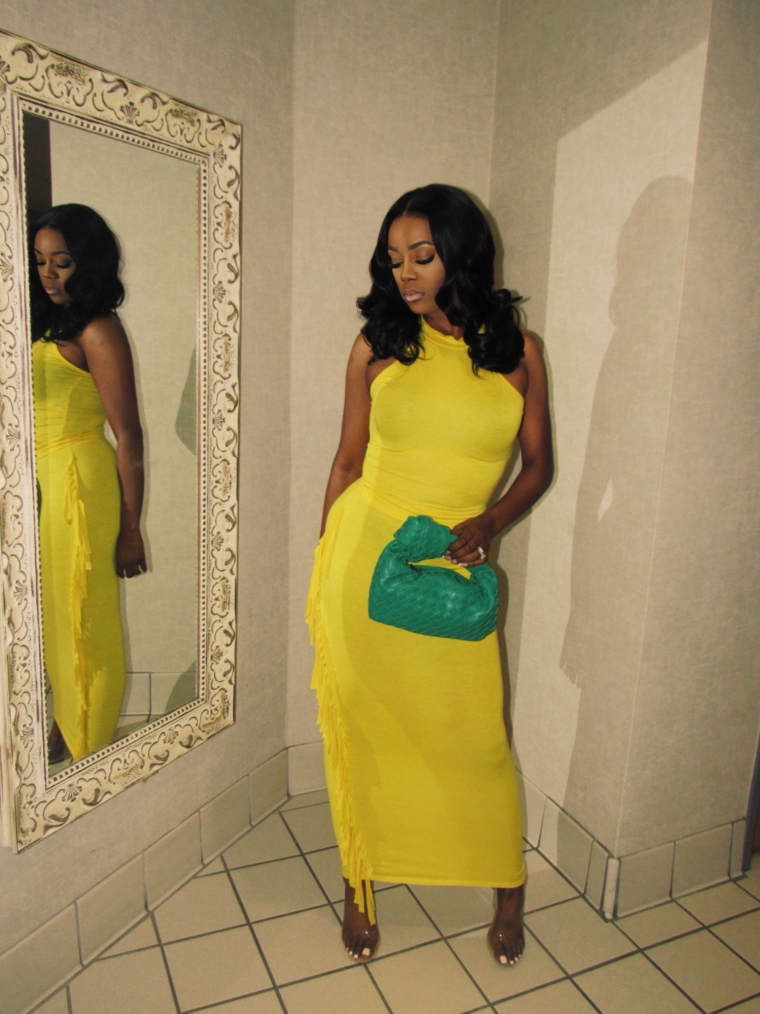 Woman in a yellow dress holding a green clutch in front of a mirror.
